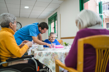 Fototapeta premium Caregiver helping an elderly woman in a blue shirt with an activity at a table in a bright common area of a nursing home