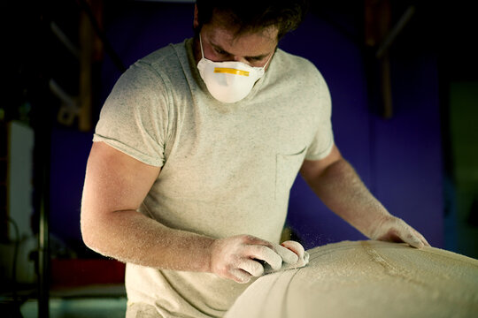 Surfboard, man and shape in workshop for manufacturing, design or polish. Surf, person and tools for sanding blank with block, smooth and production process with dust on foam for bespoke handcraft