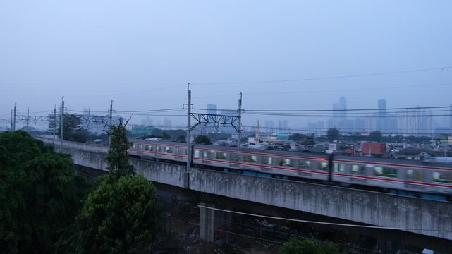 A commuter train travels on an elevated railway track with power lines above. The scene is set in an urban environment with high-rise buildings faintly visible in the background under a cloudy sky.  
