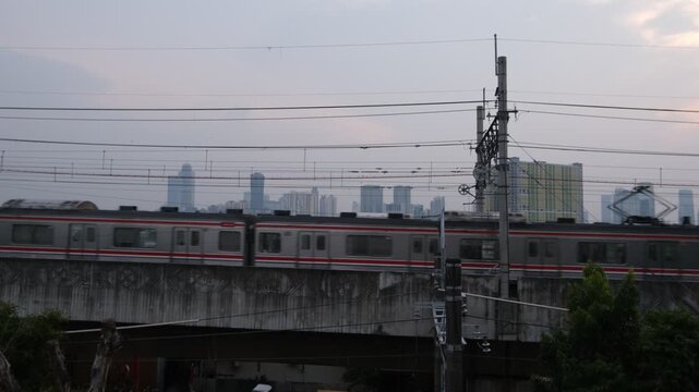 A commuter train travels on an elevated railway track with power lines above. The scene is set in an urban environment with high-rise buildings faintly visible in the background under a cloudy sky.  
