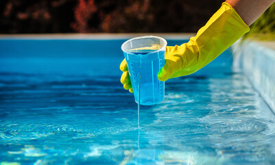 Hand in yellow protective glove pouring blue pool chemical from measuring cup into clear water for swimming pool disinfection and maintenance