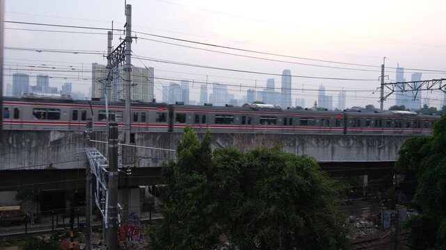 A commuter train travels on an elevated railway track with power lines above. The scene is set in an urban environment with high-rise buildings faintly visible in the background under a cloudy sky.  
