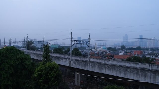 A commuter train travels on an elevated railway track with power lines above. The scene is set in an urban environment with high-rise buildings faintly visible in the background under a cloudy sky.  
