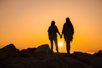 Silhouette of a mother and daughter holding hands on rocky shore at sunrise, symbolizing family love, emotional well-being, inner harmony, and a healthy lifestyle by the sea