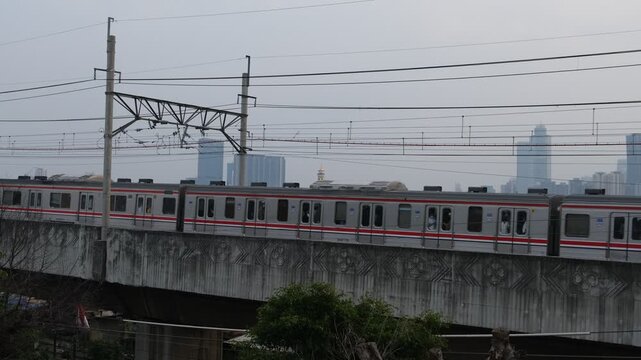 A commuter train travels on an elevated railway track with power lines above. The scene is set in an urban environment with high-rise buildings faintly visible in the background under a cloudy sky.  
