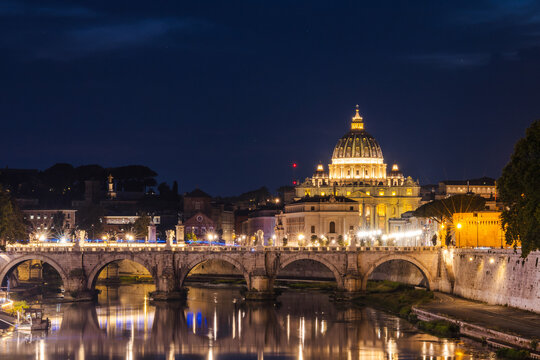 Night view of St. Peter's Basilica in Vatican City with the illuminated Ponte Sant'Angelo bridge reflected in the Tiber River in Rome