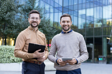 Two smiling male business colleagues stand outdoors by a modern glass office, one with a clipboard and the other with a tablet, projecting teamwork, confidence and professional partnership