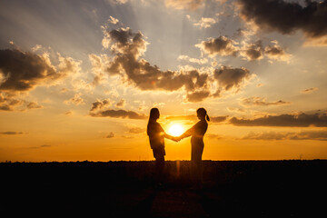 Two women standing in a field at sunset, holding hands in silhouette against the dramatic sky