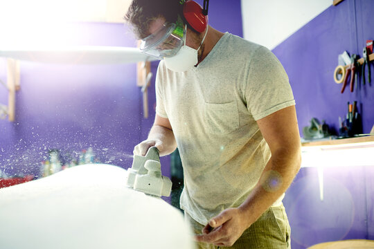 Man, tools and sanding surfboard in workshop to smooth surface, shaping process and repair work. Person, face mask or machine for diy restoration, board maintenance and polish craft to prepare finish - Powered by Adobe