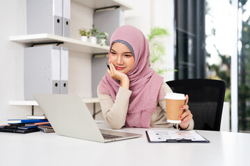 Attractive Muslim business woman in hijab working on her laptop and drinking coffee in office.