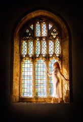 Beautiful woman with long red hair wearing a white dress, posing in the light coming through an old gothic church window