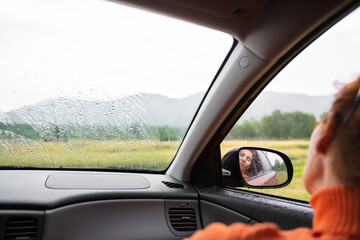 Woman relaxing in a car during the rain in the forest. Green forest and mountains in the background.