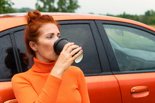 Redhead woman wearing orange sweater drinks coffee near the orange car. Green trees in the background.