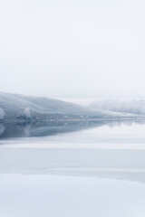 Mountains and frosted trees are reflected in the lake. Beautiful winter landscape at foggy morning