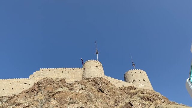 Majestic ancient fort perched high on rugged rocks under a clear blue sky in Mutrah Fort, Muscat, Oman. The stone walls and imposing circular towers showcase historical architecture and strength.