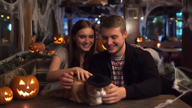 Young couple smiling while petting cat wearing tiny witch hat in halloween cat cafe with carved pumpkin lanterns and spiderweb garlands. Warm light. Concept of festive romance and pet joy