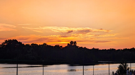 A beautiful yellow sunset over a calm river. Several clouds frame the setting sun. A row of trees outlines the forest edge on the horizon. Beautiful colorful dramatic sky with cloud.