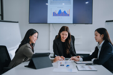Three smiling Asian businesswomen are collaborating, analyzing financial charts and data on documents and a large screen during a corporate meeting in a modern office