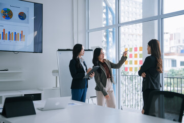 Three young businesswomen collaborating and identifying strategies during an office meeting, using a glass wall with colorful sticky notes for brainstorming sessions and project planning