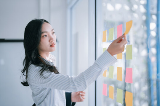 Asian businesswoman planning strategy by arranging colorful sticky notes on a glass board, actively engaging in a conceptual brainstorming session within a modern office setting