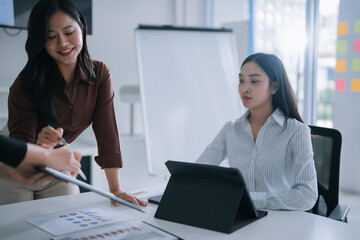 Asian businesswomen collaborating in a modern office, analyzing data and discussing financial charts on documents and a digital tablet, fostering teamwork and strategic planning