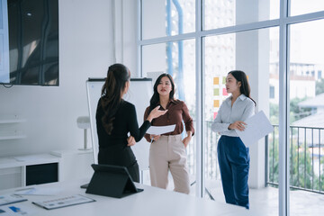 Asian businesswomen collaborating and exchanging ideas about business strategy, holding documents and sharing insights during a corporate discussion in a modern office environment