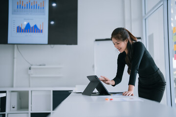 Young Asian businesswoman smiling while reviewing charts and working on a digital tablet at a modern office desk with a large screen displaying financial data in the background