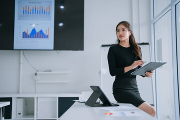 Young Asian businesswoman sits on an office desk holding a clipboard, thoughtfully reviewing financial charts on a large screen during a modern corporate meeting and strategy session