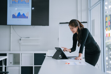 Young Asian businesswoman in a modern office analyzing financial charts and reports on a tablet during a meeting, smiling confidently while presenting data and strategy to colleagues