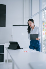 Asian businesswoman standing in a bright minimalist office, holding a clipboard and discussing an online presentation visible on a tablet, focusing on strategic planning and innovation