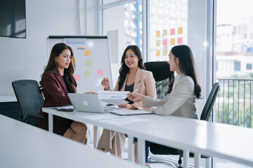 Three Asian businesswomen collaborate over documents and laptops in a modern office, analyzing charts and planning strategy during a focused team meeting for growth and finance