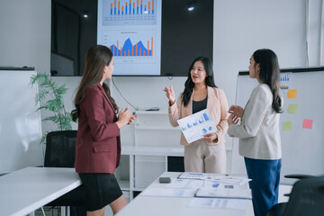 Three businesswomen actively brainstorming, analyzing charts and graphs on screen and paper, collaborating on project strategy and presenting ideas in a modern corporate office meeting room