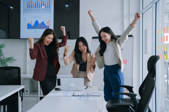Asian businesswomen cheering and jumping with arms raised in a bright modern office, celebrating team success and achievement after closing a project or reaching company goals