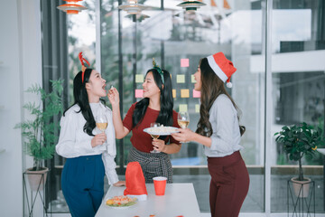Young Asian women in festive office attire laugh, share snacks and wine at a casual Christmas celebration with coworkers in a bright modern workplace, candid and joyful