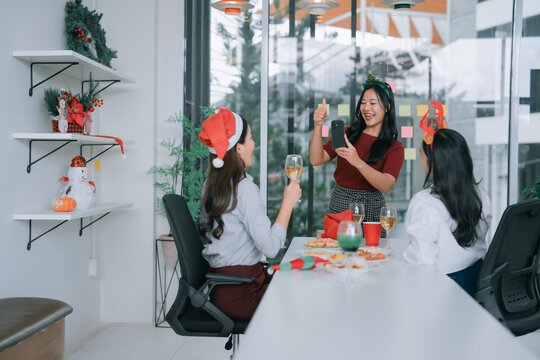 Office colleagues enjoying Christmas party in the workplace, taking selfie with a smartphone and giving thumbs up, celebrating the festive holiday season together