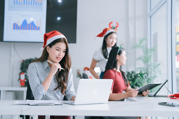 Young Asian businesswomen are diligently working at desks in a decorated office, celebrating the holiday season with Santa hats and festive accessories while reviewing documents and using laptops