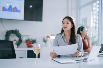 Young Asian businesswoman in a Santa hat holds documents at a decorated office desk with champagne,...