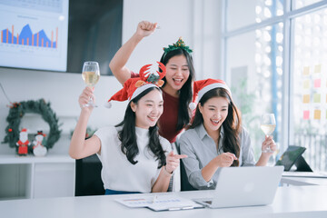 Three Asian women colleagues celebrating a festive office Christmas party. Wearing Santa hats and headbands. Holding wine glasses. And toasting while looking at a laptop receiving or making video call