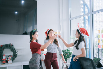 Three happy Asian women colleagues celebrating a festive Christmas office party, making a toast with drinks, enjoying their friendship and success while wearing Santa hats and holiday headbands