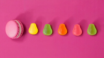 A pink macaron and colorful pear shaped candies on a pink background arranged in a horizontal line
