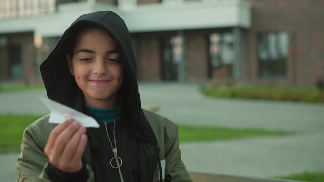 kid in hoodie collects paper jet with excitement and playful joy, smiling with dimples while preparing to fly it, capturing innocence, happiness, and creativity in casual outdoor park environment
