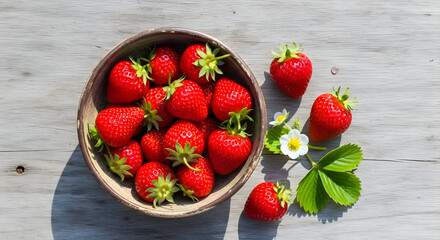 A rustic bowl overflowing with ripe, red strawberries, some scattered around with a few leaves and blossoms on a weathered wooden surface.
