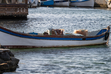 A small, traditional wooden fishing boat, painted white with a blue trim, is moored in rippling water with fishing gear visible inside.