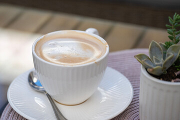 A ribbed white ceramic cup holds a foamy cappuccino or latte, sitting on a saucer next to a small succulent plant with a spoon resting below the rim.