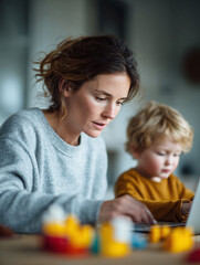 Obraz premium Mother multitasking while working on a laptop with her young child nearby. Depicts modern family life, worklife balance, and parental responsibilities. Home office concept.