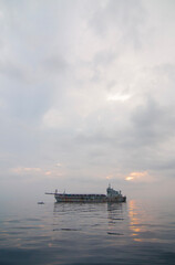 Obraz premium A large cargo ship is anchored on the exceptionally calm waters of Ternate, Indonesia, at dawn or dusk. A peaceful and minimalist scene with the sea reflecting the soft, cloudy sky.