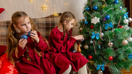 Little sisters, beautiful child girls in red dresses are decorating a Christmas tree in living room. winter New Year's concept.