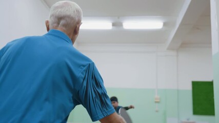 Back view of senior trainer in blue shirt serving tennis ball across indoor table while focused trainee prepares to return shot during intense training session in brightly lit sports hall - Powered by Adobe