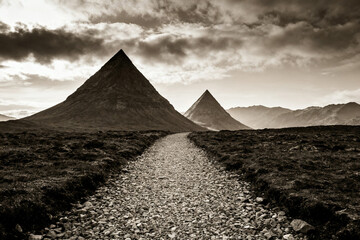 A gravel path leads toward two prominent pyramidal mountains under a dramatic cloudy sky.