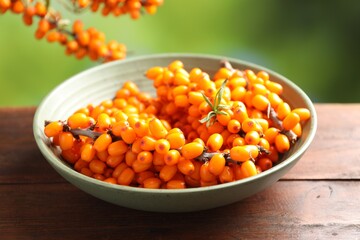 Ripe sea buckthorn berries and leaves in bowl on wooden table against blurred green background, closeup